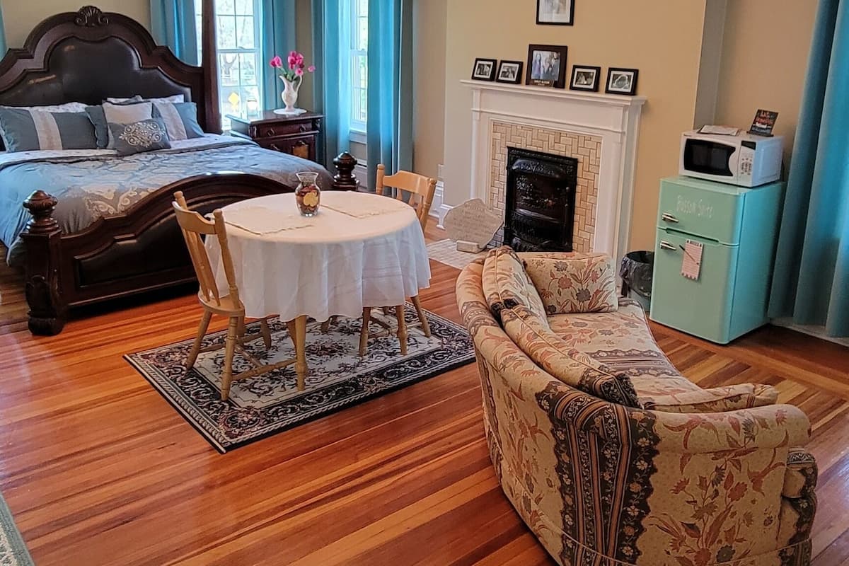A spacious living room with a large bed, a fireplace, a dining table, and a patterned sofa.