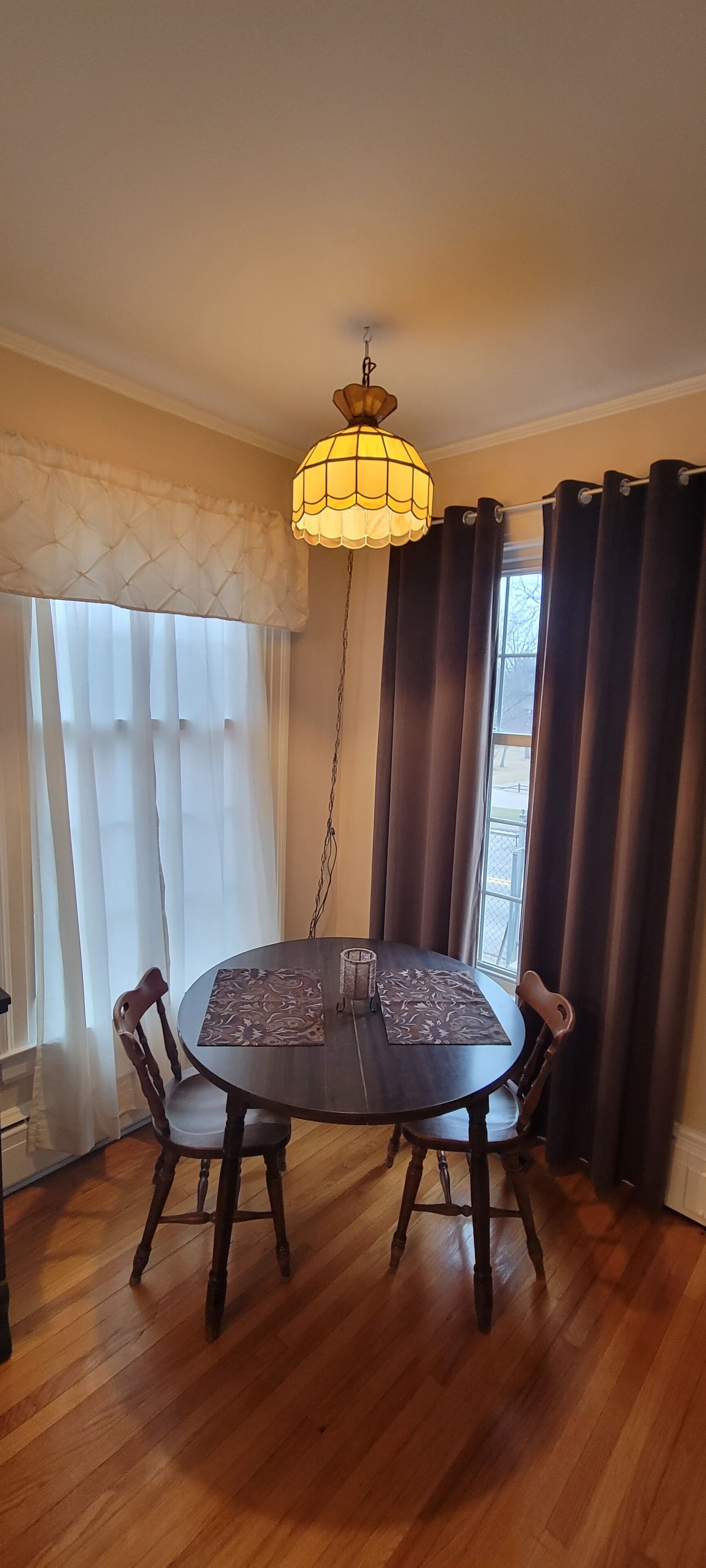 A dining area with a round table, four chairs, a hanging light fixture, and a window with curtains.