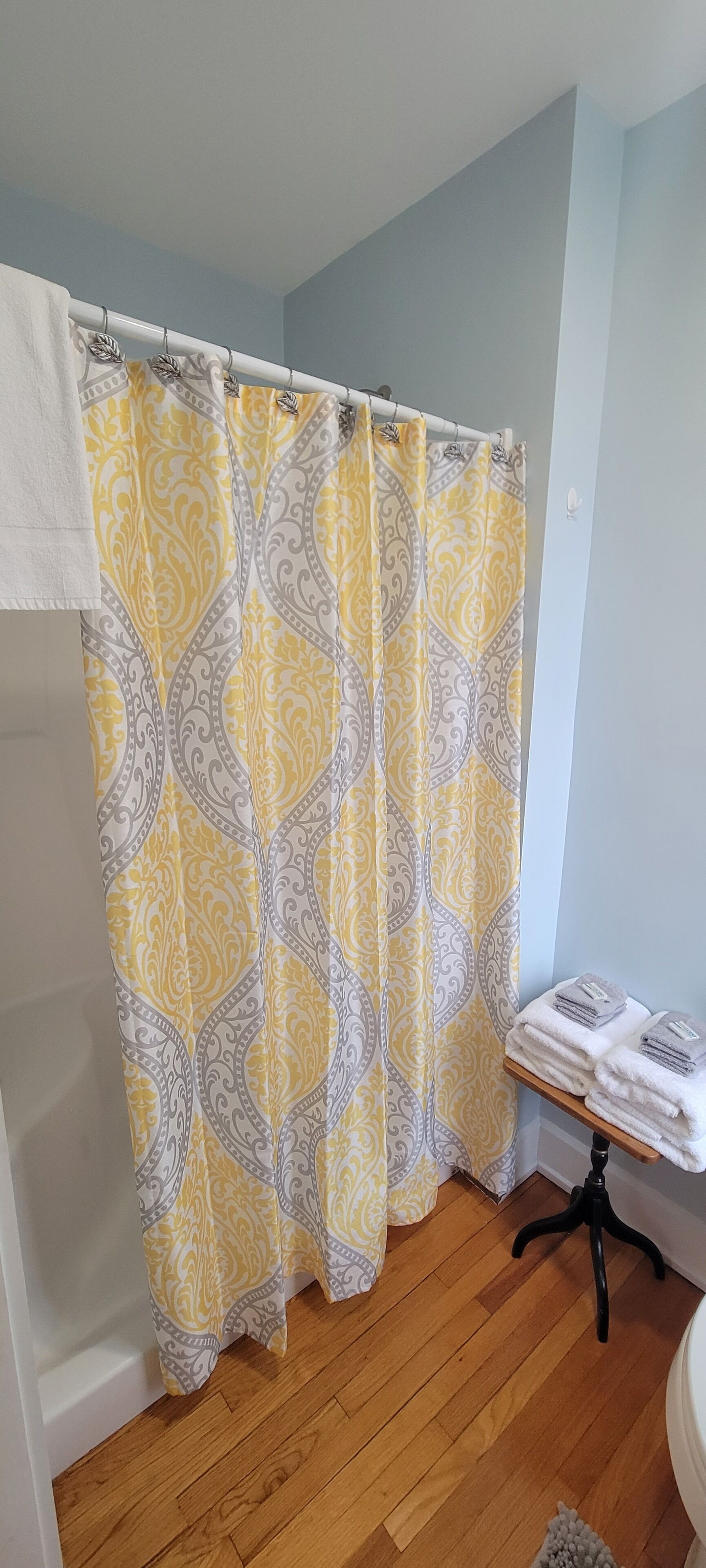 A bathroom with a shower curtain featuring a yellow and gray paisley pattern, a wooden floor, and a small table with folded towels.