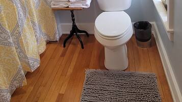A bathroom with a toilet, a small table with towels, a framed picture on the wall, a patterned shower curtain, and a rug on the floor.
