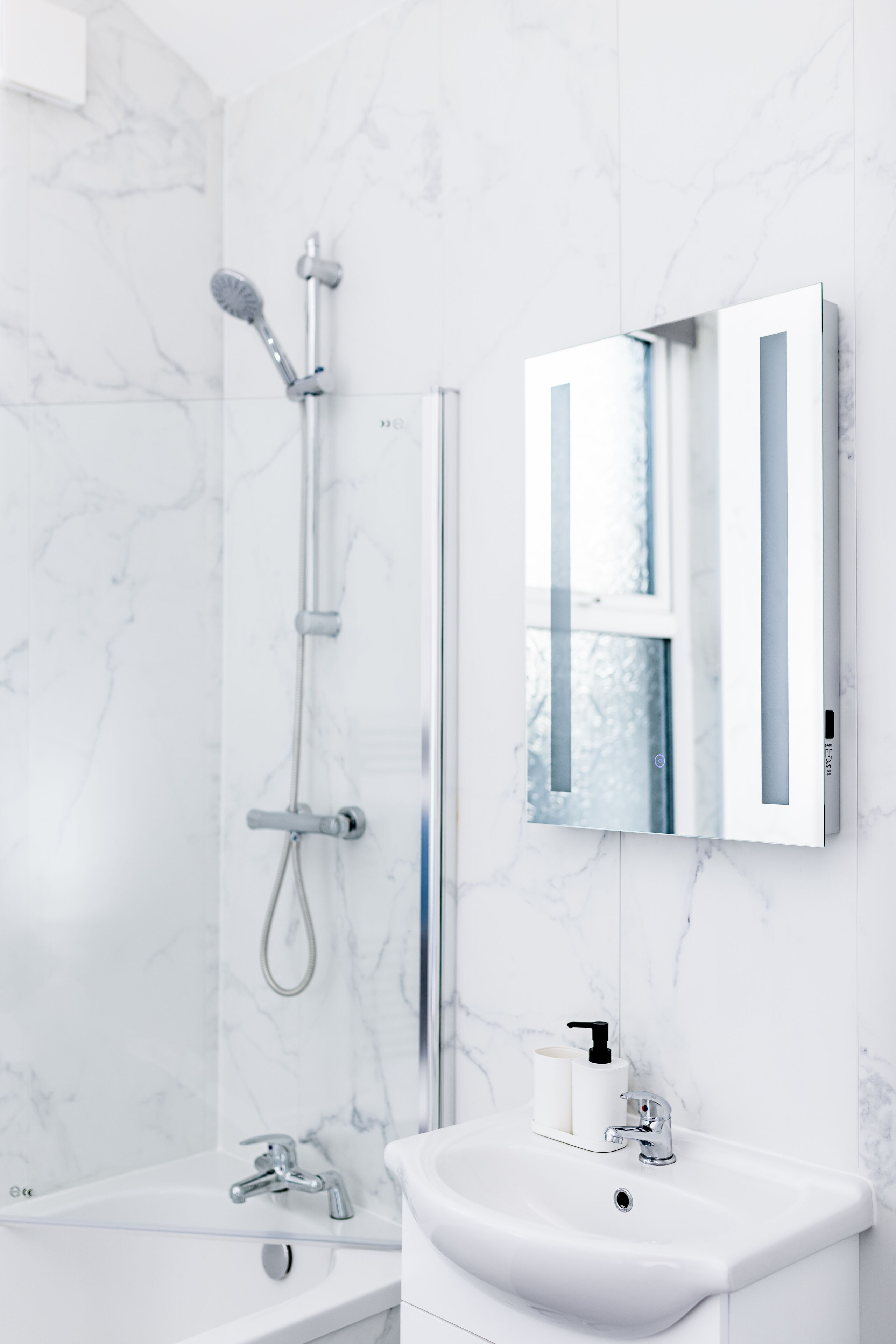 A modern bathroom with a white marble wall, a glass shower enclosure, a white sink, and a mirror with a white frame.