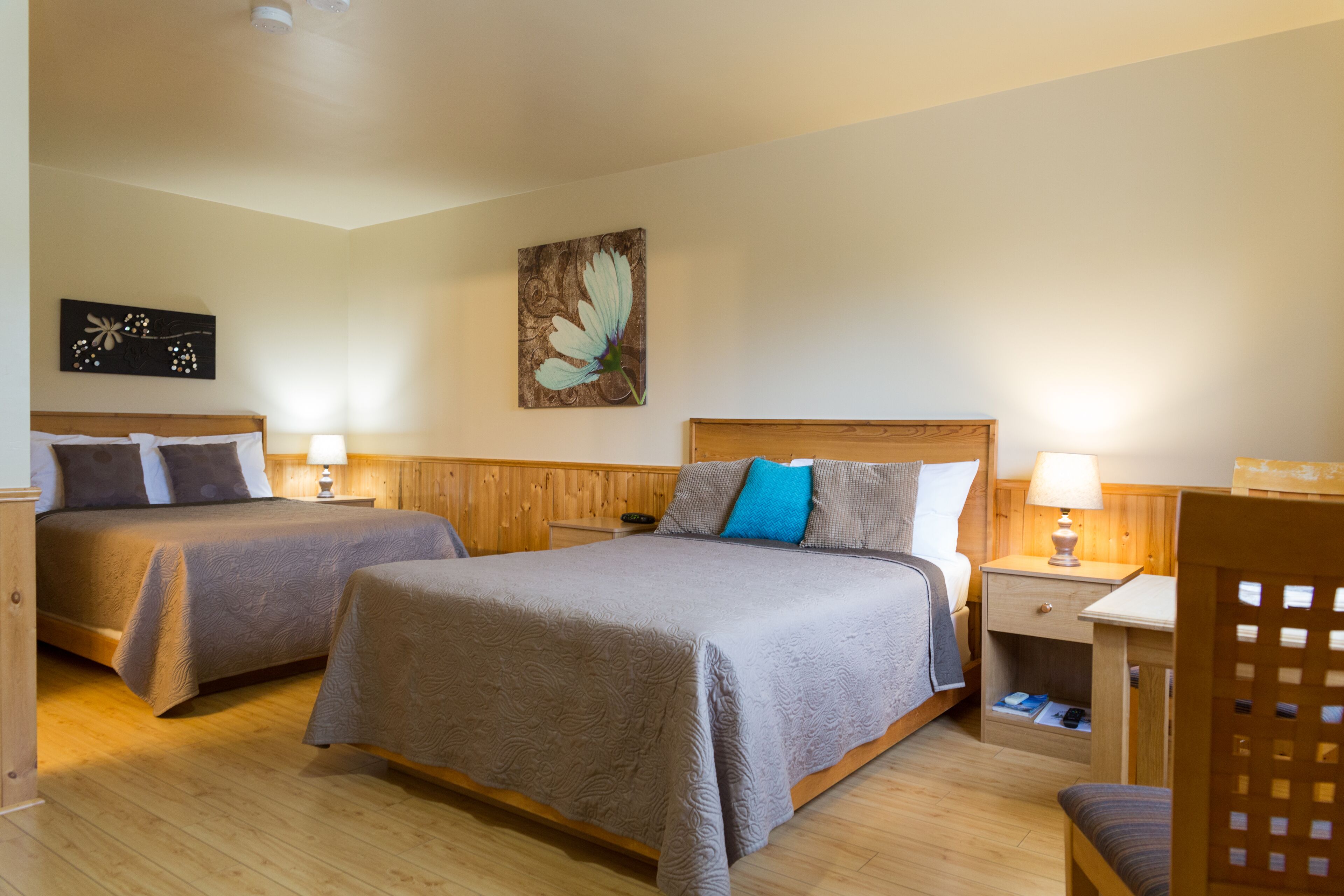 A hotel room with two beds, wooden flooring, a nightstand with a lamp, and a wall painting.