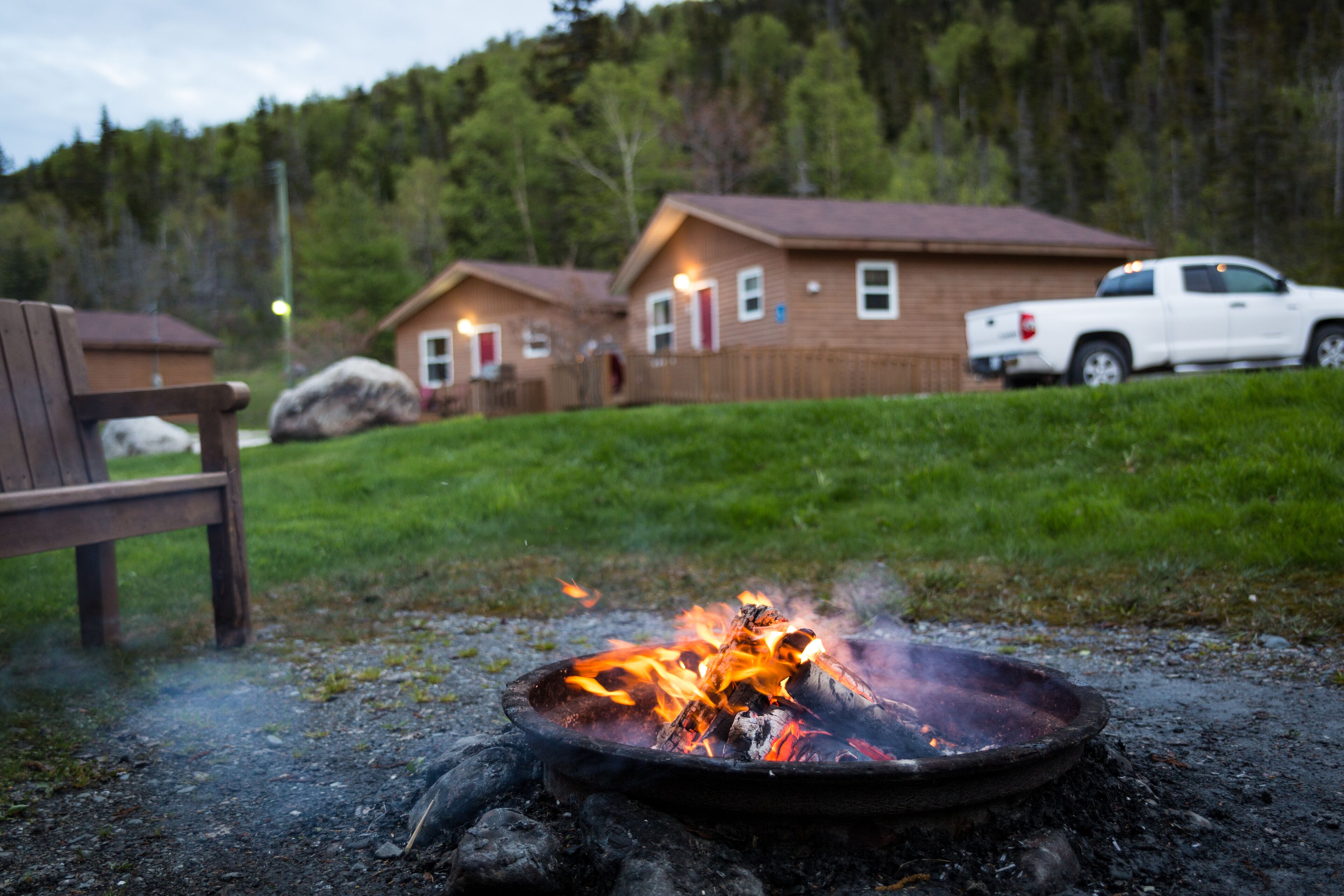 A campfire with a metal bowl, a wooden bench, and a white pickup truck parked near cabins in a wooded area.