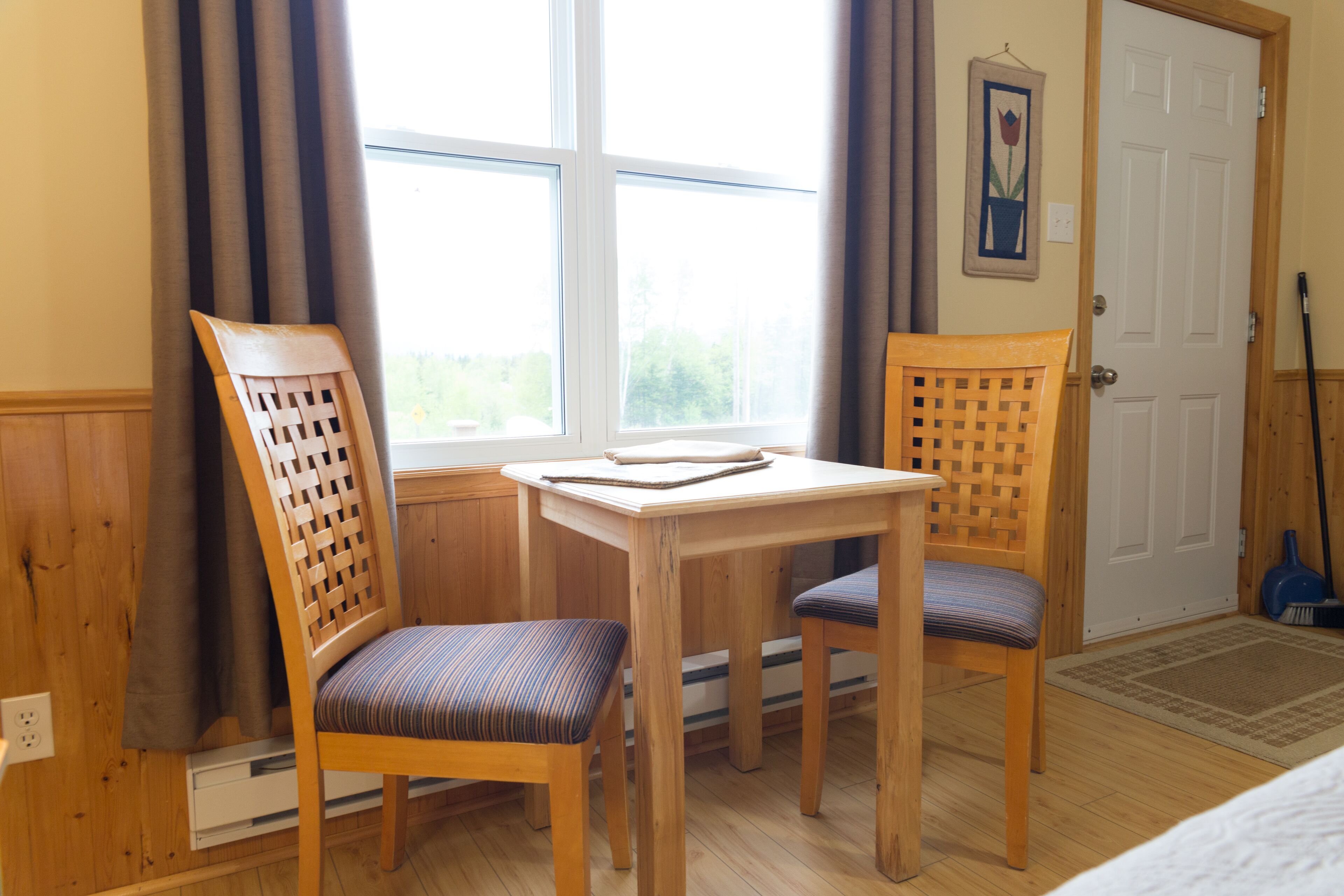Una habitación con una mesa de madera y dos sillas, una ventana con cortinas, una puerta y un cuadro en la pared.