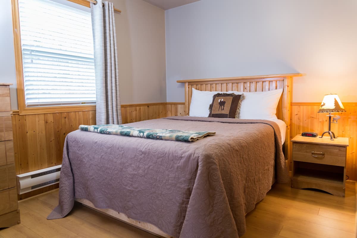 A bedroom with a wooden headboard, a bed with a purple blanket, a nightstand with a lamp, and a window with blinds.