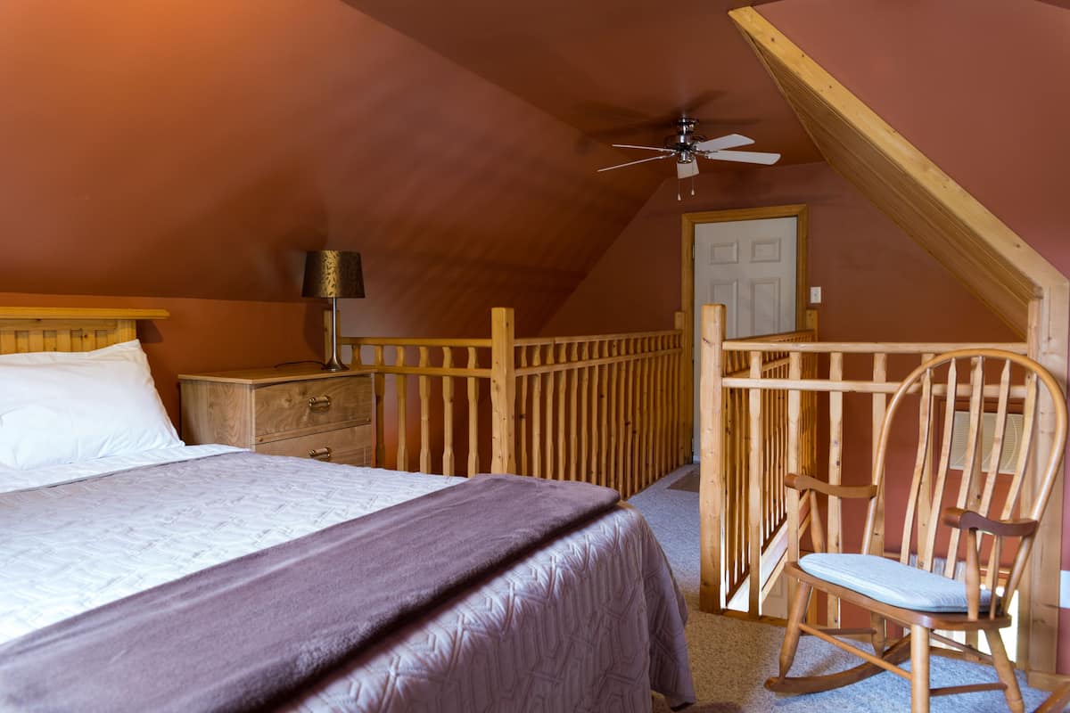 A bedroom with a bed, a wooden rocking chair, a nightstand, a lamp, and a ceiling fan.