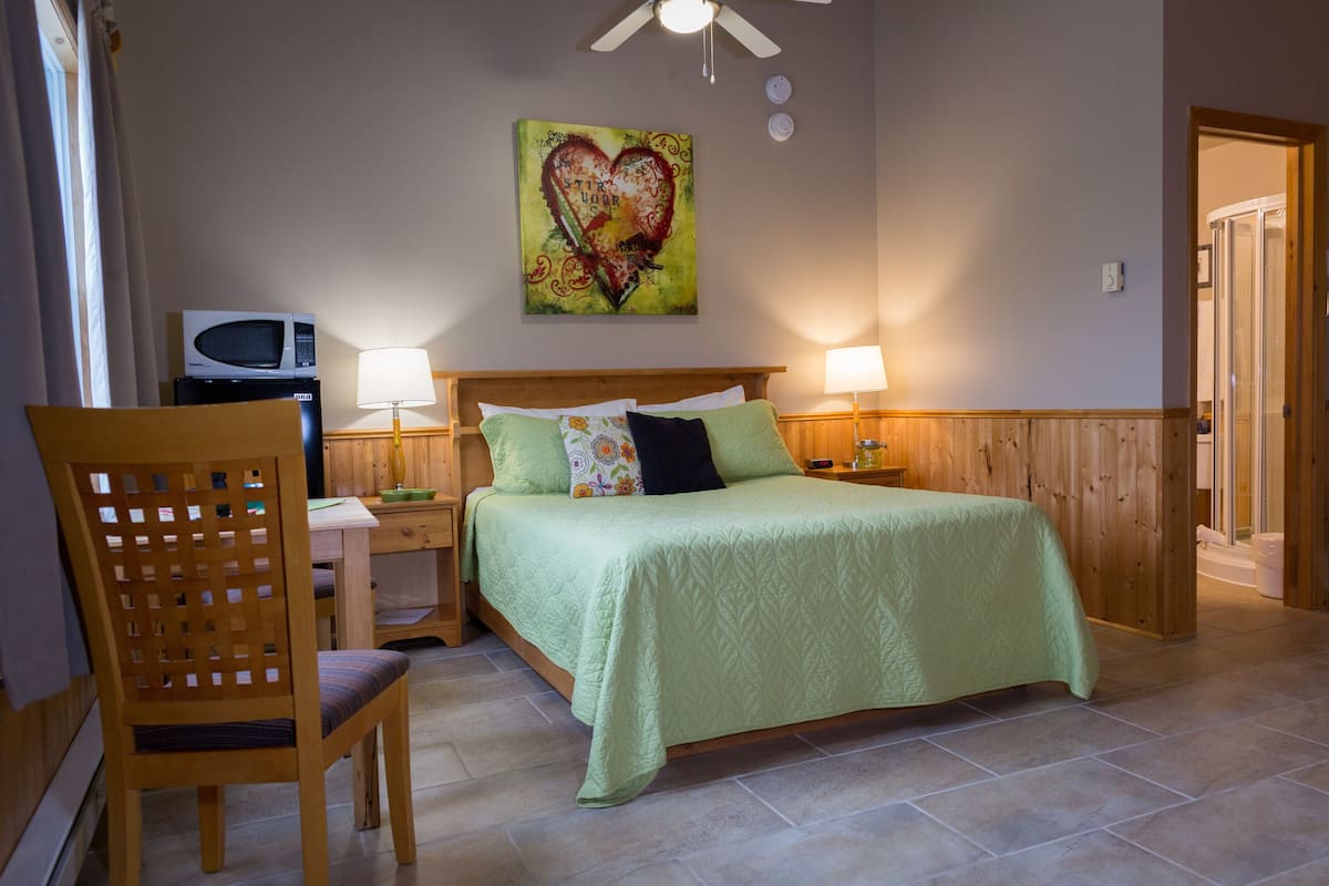 A bedroom with a bed, two bedside lamps, a wooden headboard, a chair, a microwave, and a heart-themed artwork on the wall.