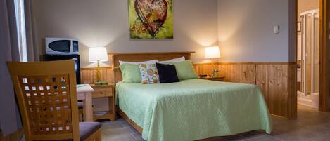 A bedroom with a bed, two bedside lamps, a wooden headboard, a chair, a microwave, and a heart-themed artwork on the wall.