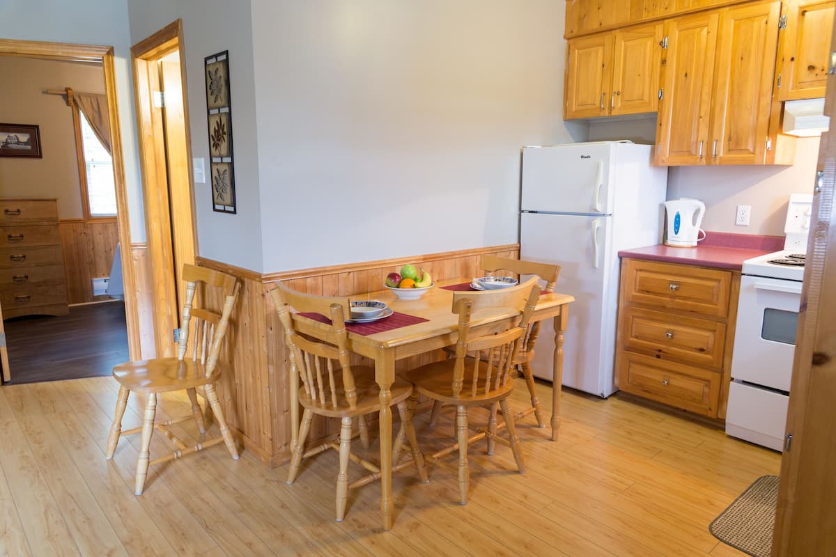 A wooden dining table with four chairs, a refrigerator, a coffee maker, and a fruit bowl on the table.