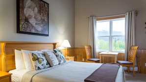 A neatly arranged bedroom with a large bed, a bedside table with a lamp, a small desk, and a chair. There is a window with curtains and a view of greenery outside. - Middle Brook Cottages & Chalets (Glenburnie-Birchy Head-Shoal Brook)