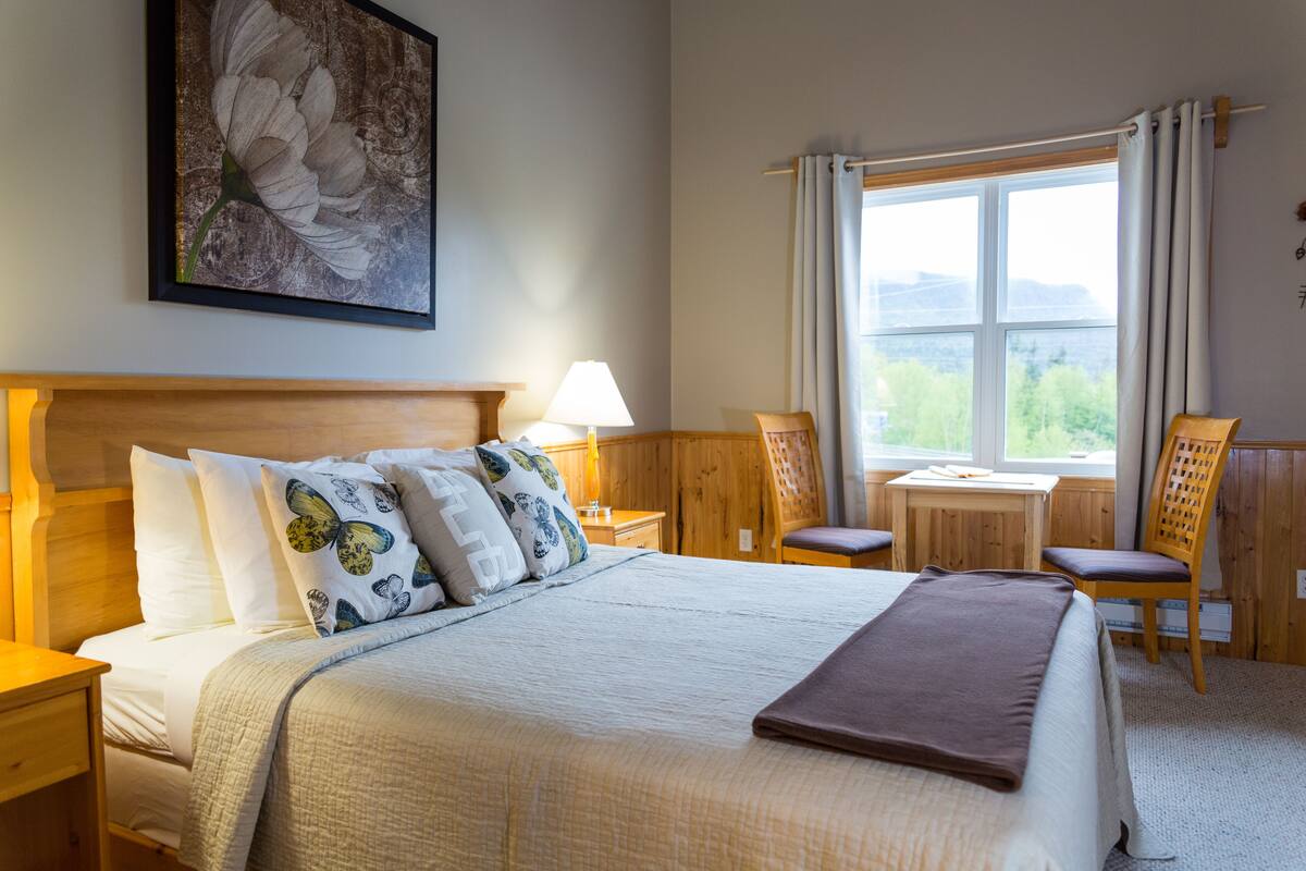 A neatly arranged bedroom with a large bed, a bedside table with a lamp, a small desk, and a chair. There is a window with curtains and a view of greenery outside.