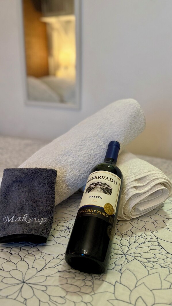 Una botella de vino Malbec, una toalla blanca doblada y una toalla azul oscuro doblada sobre una cama con cubrecama estampado.