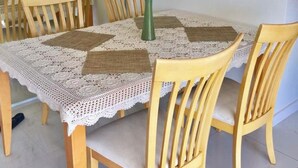 Uma área de refeições com uma mesa redonda coberta por um tecido de renda, rodeada por cadeiras de madeira com assentos acolchoados. Um vaso com uma única rosa branca está sobre a mesa.