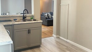 A modern kitchen with a marble countertop, a sink, and a built-in cabinet. There is a hallway leading to a living area with a couch and a door. - Large group & couples retreat (Pineville)
