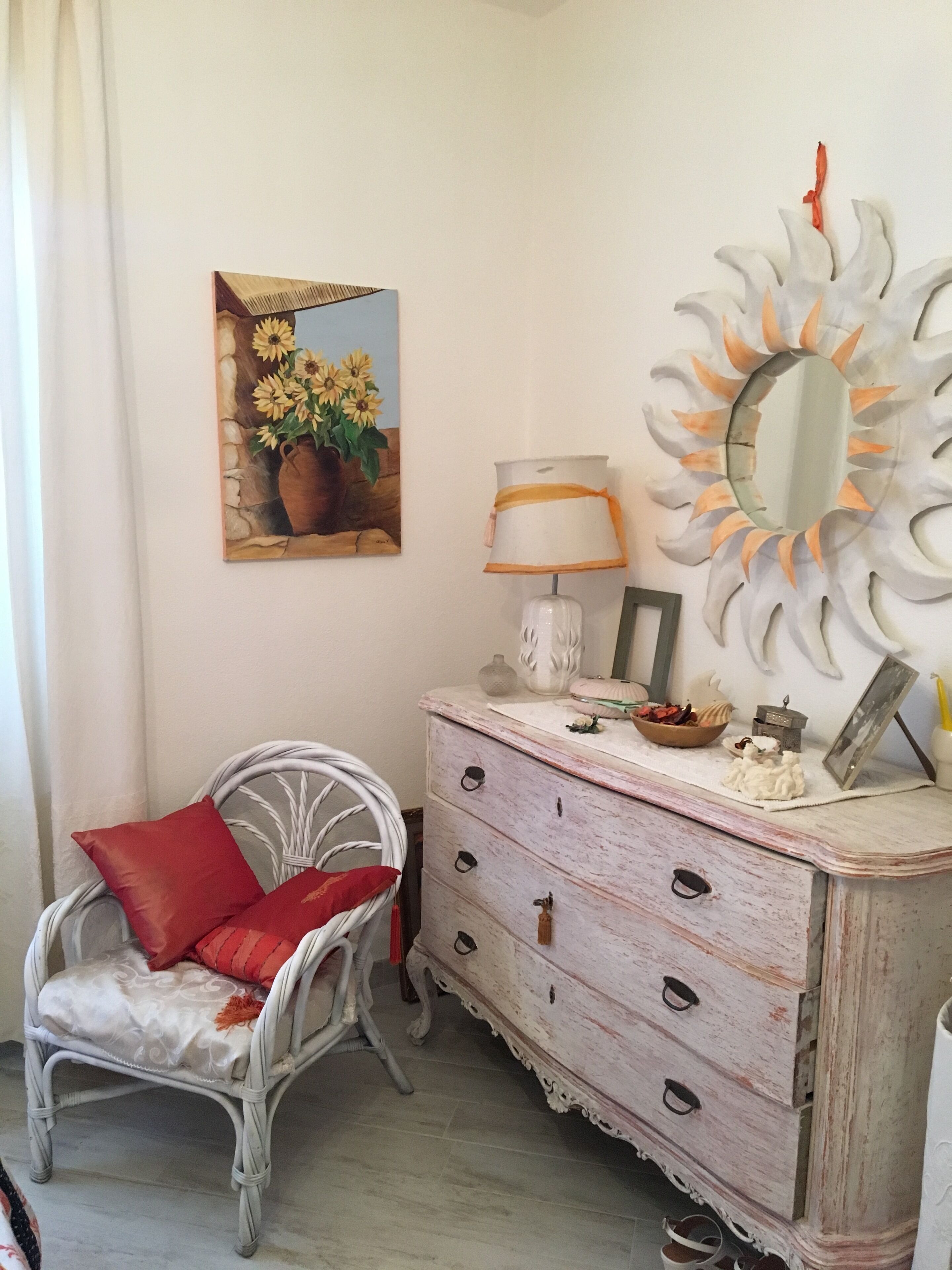 A room with a white wicker chair, a white dresser with drawers, a mirror with a sun-like frame, and a painting of sunflowers on the wall.