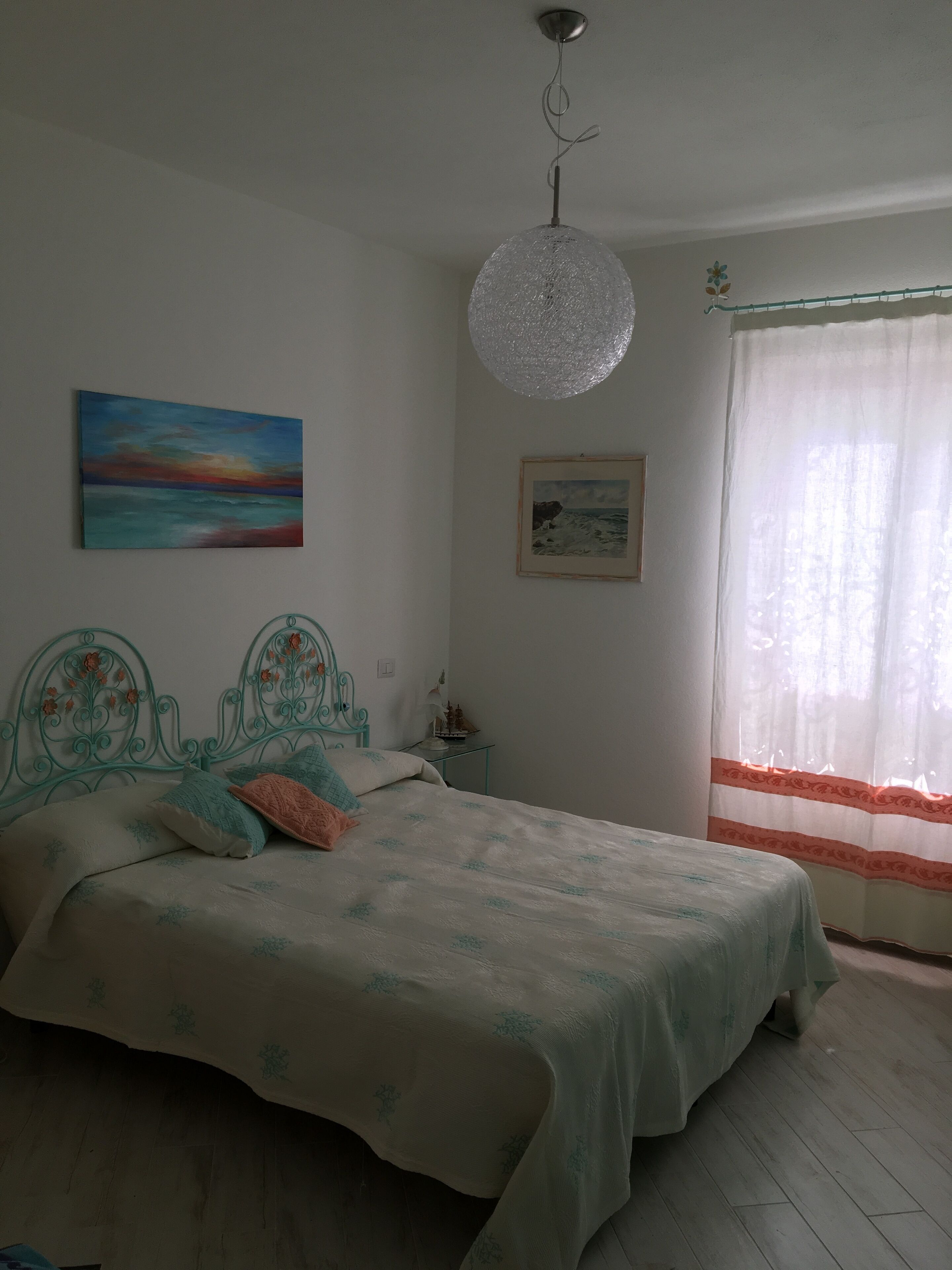 A bedroom with a bed, a hanging light fixture, a painting, a framed picture, and a window with curtains.