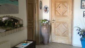 Una stanza con una porta in legno, un vaso grande con fiori, un tavolino con una ciotola e un quadro appeso al muro.