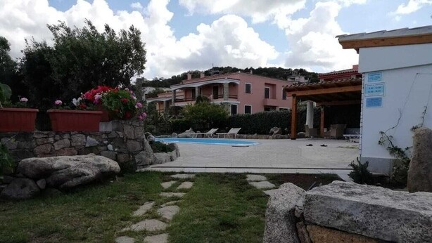Area piscina con sedie a sdraio, un vialetto in pietra e un edificio con tetto rosso sullo sfondo.