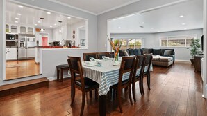 Amplia sala de estar con una mesa de comedor dispuesta para seis personas, una cocina al fondo y un gran ventanal que proporciona luz natural.