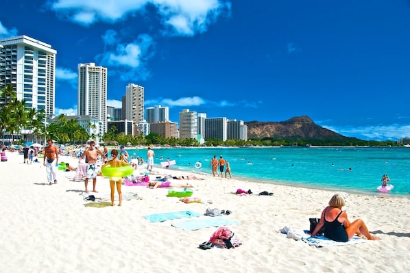 A beach with people sunbathing and swimming, palm trees, and a city skyline in the background. - 265 steps to the King Kamehameha statue on Waikiki and soft white sandy beaches (Honolulu)