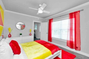 Un dormitorio con una cama, un ventilador de techo, una ventana con cortinas rojas y un espejo en la pared.