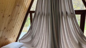 Une pièce chaleureuse avec un plafond en pente, des murs en bois et une grande fenêtre avec des rideaux. On y trouve deux lits avec du linge de lit blanc et une serviette enroulée sur l’un des lits.