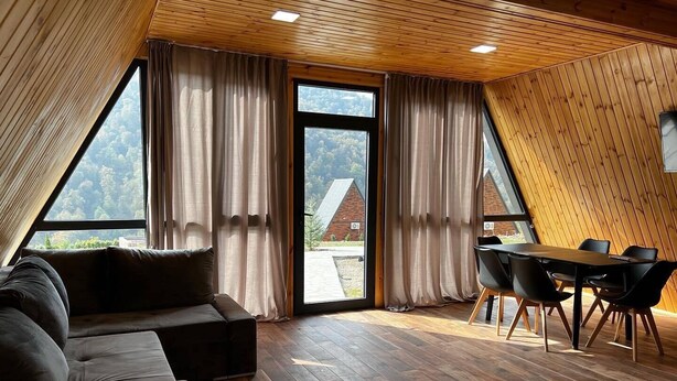 Une pièce chaleureuse avec un plafond en bois, un canapé, une table à manger avec des chaises et de grandes fenêtres avec des rideaux.