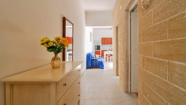 A hallway with a wooden dresser and a vase of flowers. There is a kitchen area with cabinets and a dining table in the background.