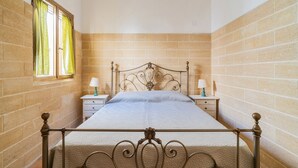 A bedroom with a metal bed frame, a window with curtains, a bedside table with a lamp, and a brick wall.