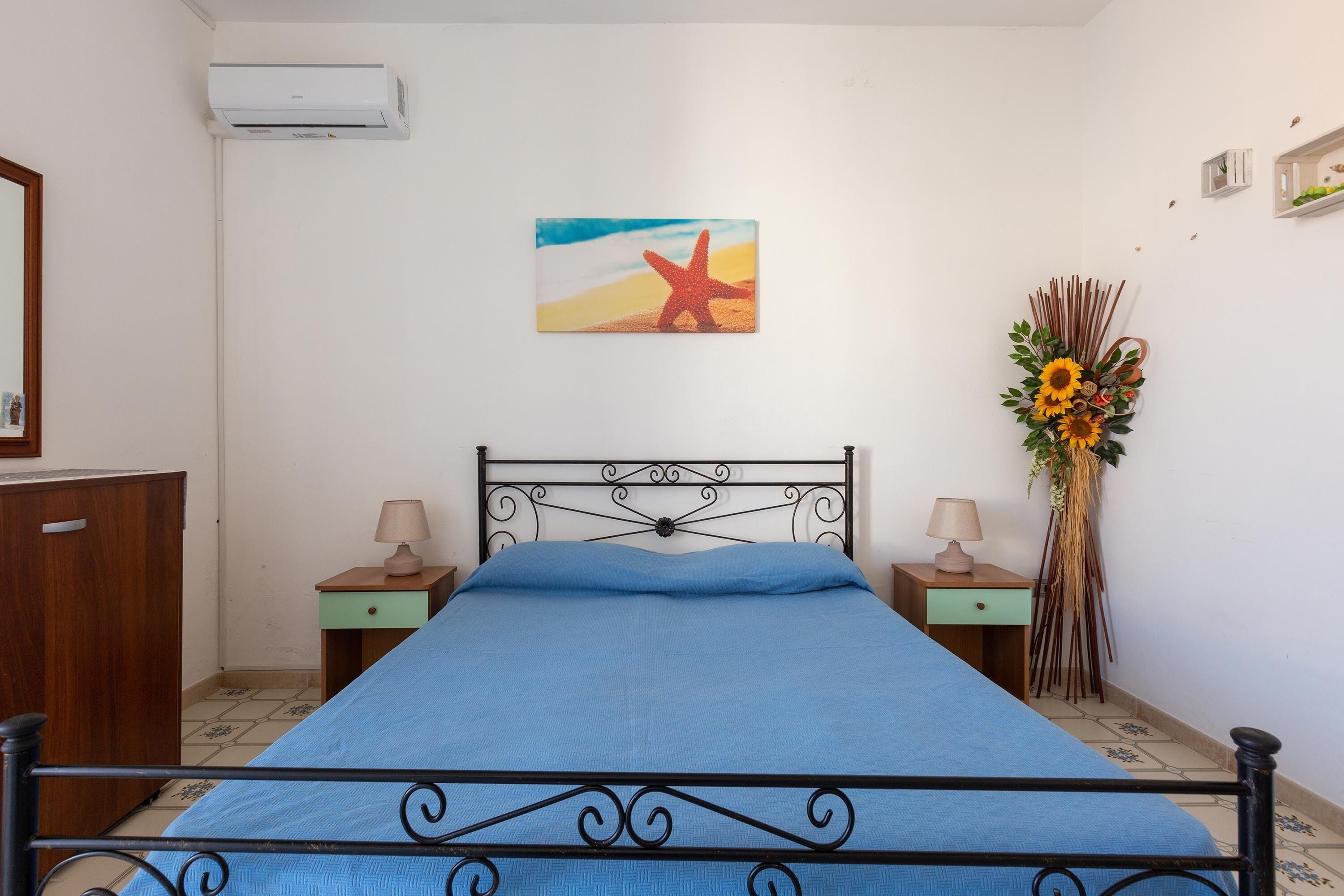 Ein Schlafzimmer mit einem Bett, zwei Nachttischen, einer Wand-Klimaanlage, einem gerahmten Bild und einer Zimmerpflanze.