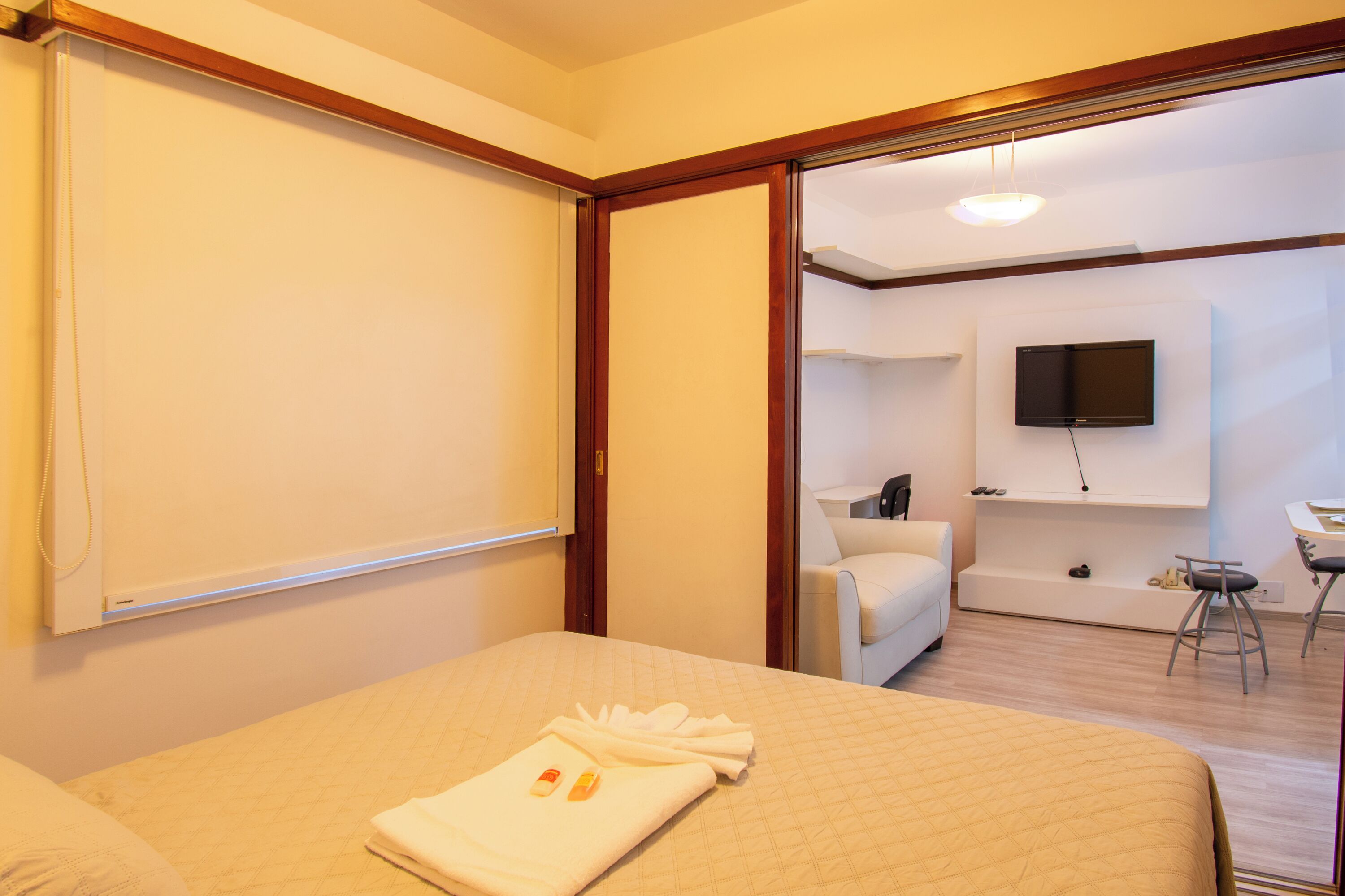 A hotel room with a bed, a TV mounted on the wall, a seating area with a chair and a small table, and a bathroom visible through an open door.