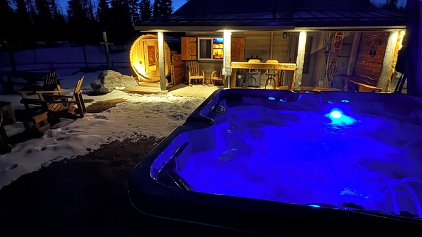 Un jacuzzi exterior con calefacción, con una terraza y sillas de madera, una cabaña de madera con luz en su interior y un paisaje nevado.