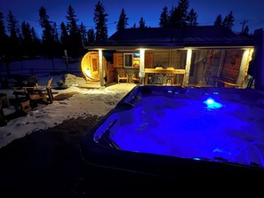 Un bain à remous extérieur chauffé, doté d’une terrasse en bois et de chaises, un chalet en bois dont l’intérieur est éclairé, et un paysage enneigé.