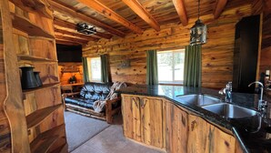 Interior de una cabaña de troncos con sala de estar, un sofá, chimenea y cocina con pileta.