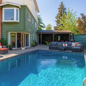 A pool area with a built-in seating area, a covered patio, and a house with green siding. - Hidden Oasis in Victoria, BC (Victoria)