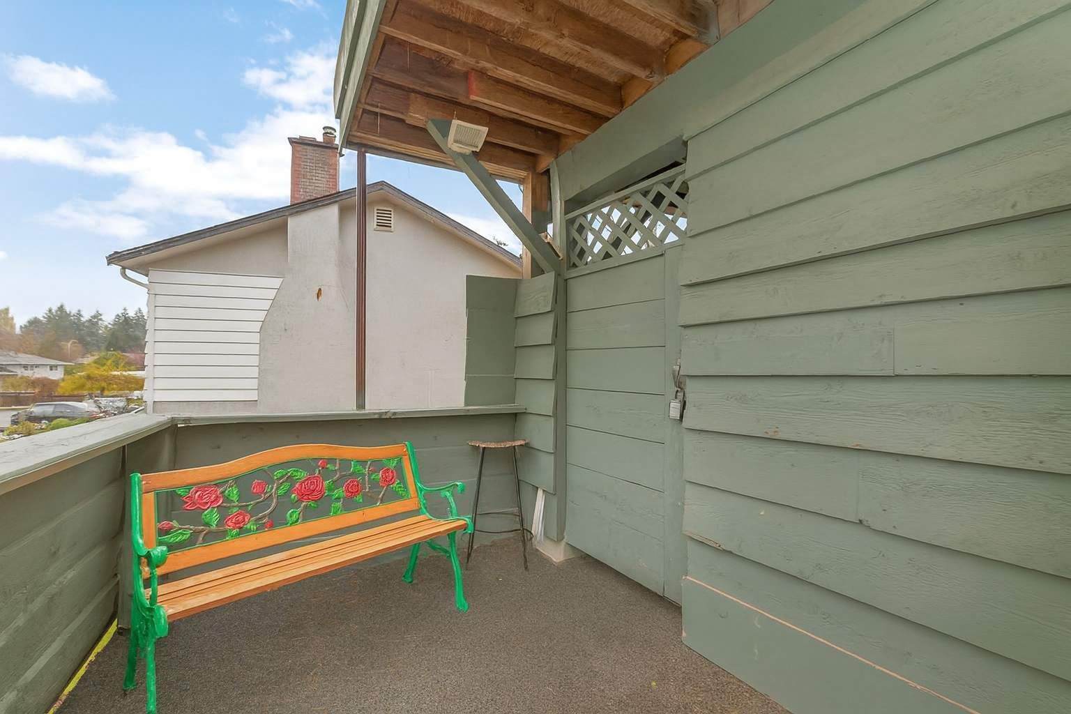 A covered outdoor area with a wooden bench featuring a floral design, a small table, and a green wooden wall.