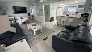 Sala de estar com sofá de couro preto, mesa de centro branca e televisão. Há uma área de cozinha com geladeira, fogão e banquetas de bar.