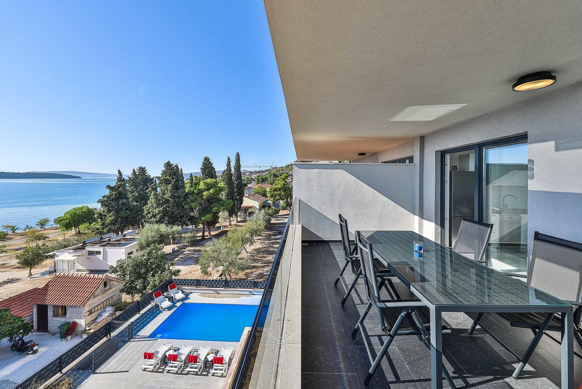 A balcony with a glass table and chairs overlooking a swimming pool and a scenic view of the sea and trees.