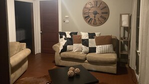 Een woonkamer met een beige bank, een bijzettafeltje met decoratieve ballen, een grote houten klok en een plafondventilator.