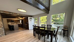 A spacious dining area with a wooden table and chairs, a kitchen in the background, and large windows offering a view of greenery.