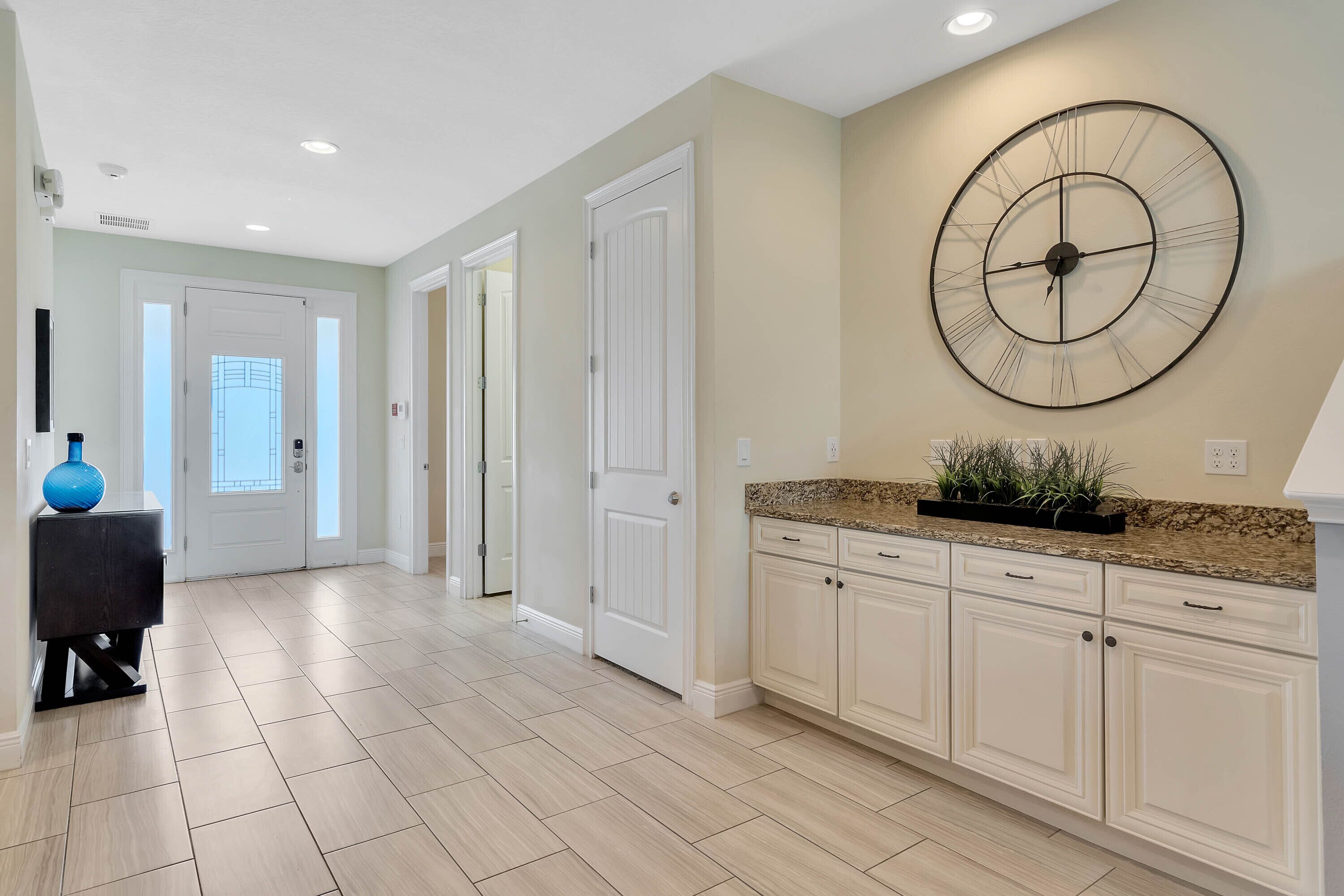 A spacious kitchen with white cabinets, a large wall clock, and a granite countertop.