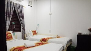 A hotel room with two beds, each with a white bedspread and a red and yellow patterned blanket. There is a window with curtains, a bedside table with a lamp, and a green trash bin. - Chitlatda Guesthouse (Luang Prabang)