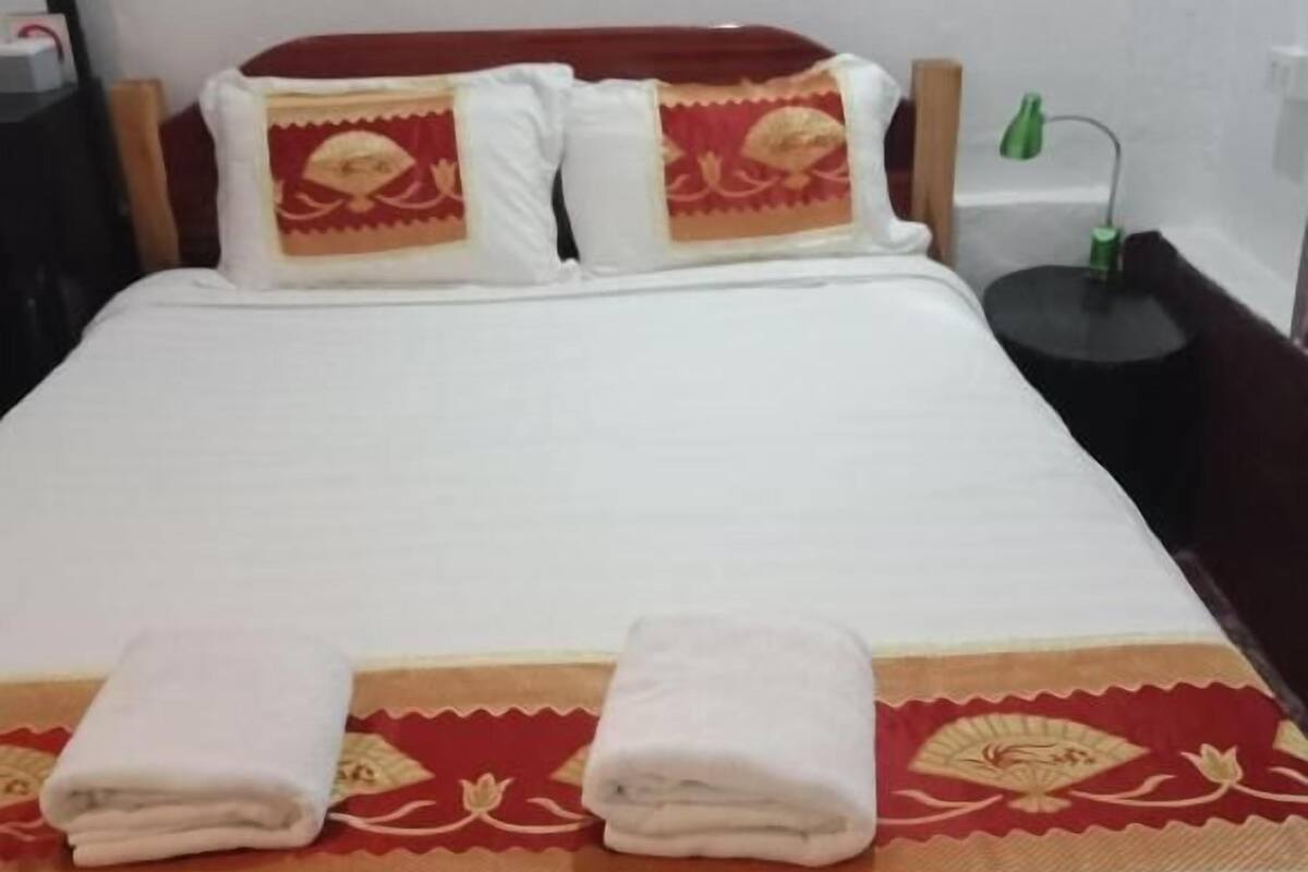 Una cama bien tendida con una colcha blanca y dos almohadas, un corredor decorativo rojo y dorado, y dos toallas dobladas sobre la cama.
