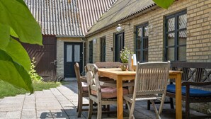 Een houten tafel met stoelen en een bloempot voor een gebouw met bakstenen muren en een pannendak.
