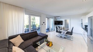 Ein modernes Wohnzimmer mit einer dunkelgrauen Couch, einem Glastisch und einem Essbereich mit weißem Tisch und Stühlen. Es gibt eine Küchenzeile mit Kühlschrank und einem Fernseher, der über der Arbeitsplatte angebracht ist.