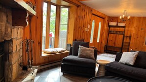 A cozy living room with a stone fireplace, wooden walls, and a large window with curtains.