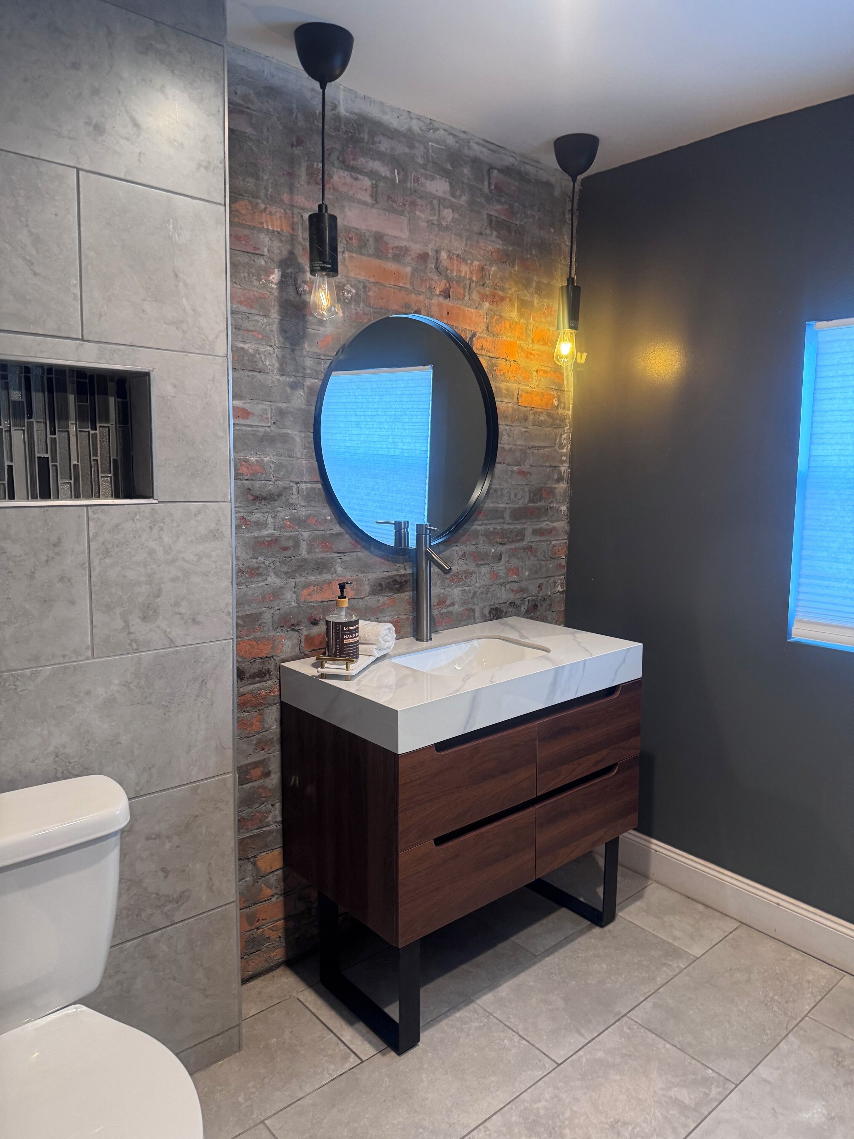 Uma casa de banho com design moderno, destacando um espelho redondo, uma pia branca e um móvel de vaidade em madeira escura.