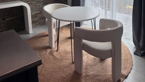 A modern living room with a brick wall, a white round table, two white chairs, a dark wooden table, and a large window with curtains.