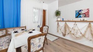 Ein Essbereich mit Tisch für zwei Personen, Stühlen mit floralem Bezug, einer Topfpflanze auf einem Regal und einer Wand mit Netzdekor und einem Gemälde.