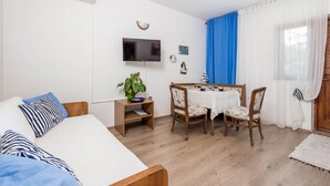 Ein Zimmer mit einem Bett, einem Fernseher, einem Esstisch mit Stühlen, einer Pflanze und einem kleinen Regal.